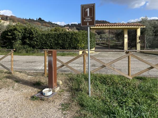 Refreshment area, marked by a touristic sign, with water fountain, at break in the fence.